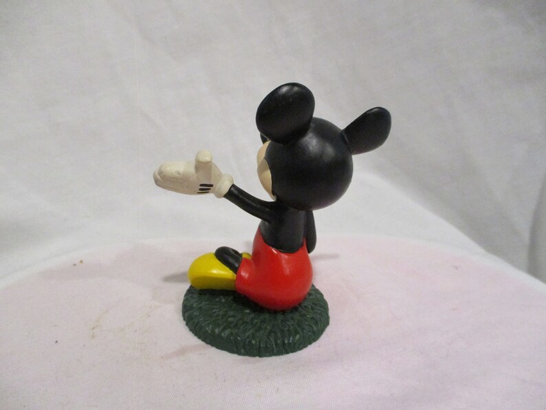 Puede incluir: Una peque&ntilde;a figura de Mickey Mouse de color negro y rojo sentada sobre una base verde. Mickey lleva sus caracter&iacute;sticos pantalones cortos rojos y zapatos amarillos. Est&aacute; saludando con la mano derecha.