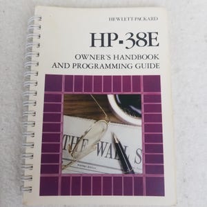 May include: A spiral-bound HP-38E Owner's Handbook and Programming Guide. The cover features the text "HP-38E" and "Owner's Handbook and Programming Guide". The cover has a purple border with an image of a newspaper, glasses, and a pen.