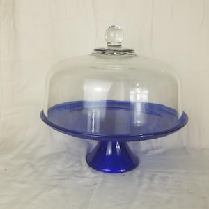 May include: A cobalt blue glass cake stand with a clear glass dome. The stand has a round base and a pedestal. The dome has a small, round handle on top. The stand is approximately 12 inches in diameter.
