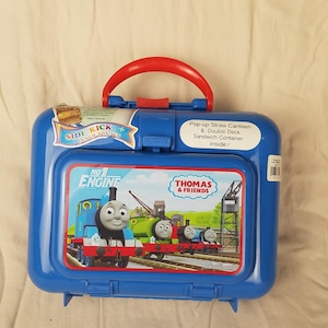 May include: A blue plastic lunch box with a red handle featuring Thomas the Tank Engine and friends. The lunch box has a pop-up straw canteen and double-deck sandwich container inside. The text "SIDE KICK" and "THOMAS & FRIENDS" are visible.