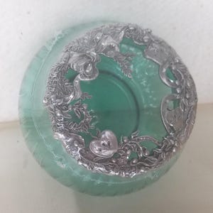 May include: A green glass jar with a decorative silver-colored metal rim. The rim features a floral and heart design. The jar is round and has a slightly textured surface. The jar is likely used for storing small items or as a decorative piece.