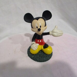 Puede incluir: Una figura de pl&aacute;stico negra y roja de Mickey Mouse sentado en una base verde. Mickey lleva zapatos amarillos y tiene un guante blanco en su mano derecha.
