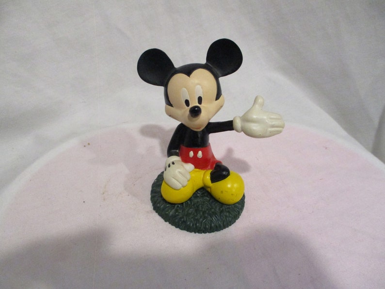 Puede incluir: Una figura de pl&aacute;stico negra y roja de Mickey Mouse sentado en una base verde. Mickey lleva zapatos amarillos y tiene un guante blanco en su mano derecha.