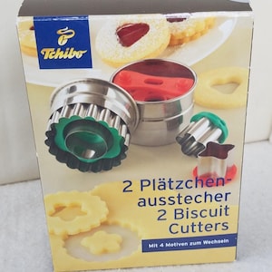 May include: A box of cookie cutters with four interchangeable designs. The box features a red and white logo with the text "Tchibo" and the text "2 Plätzchen-ausstecher 2 Biscuit Cutters Mit 4 Motiven zum Wechseln".
