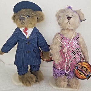 Könnte beinhalten: Zwei Teddybären im Vintage-Stil, gekleidet in Kleidung aus den 1920er Jahren. Der männliche Bär trägt einen blauen Nadelstreifenanzug, eine rote Krawatte und einen Hut. Der weibliche Bär trägt ein pinkfarbenes Paillettenkleid mit violettem Saum und eine Perlenkette. Das Etikett lautet "Daisy  Russ Berrie  1920's 20th Century Collectibles".
