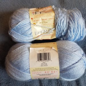 Könnte beinhalten: Zwei Knäuel hellblaues Bernat Baby Garn mit einem Gewicht von jeweils 140 Gramm. Die Etiketten zeigen den Markennamen, Pflegehinweise, einen Strichcode und die Farbbezeichnung Pale Blue. Das Garn besteht aus 100% Acryl und ist maschinenwaschbar.