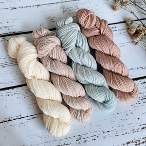May include: Four skeins of yarn in cream, light pink, seafoam green, and peach. The yarn is arranged diagonally on a white wooden surface. Dried flowers are in the upper right corner.