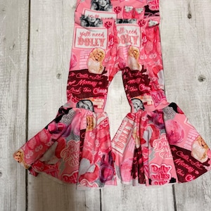May include: Pink bell-bottom pants with a repeating pattern featuring images of a celebrity, text that reads "Y'all need Dolly", and other graphics. The pants have a high waist and flared legs.