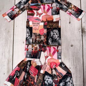 May include: A one-piece jumpsuit with long sleeves and flared legs. The fabric features a collage of images, including a star with a butterfly, a trailer park sign, and portraits. The color palette includes pink, black, and white.