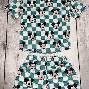 Puede incluir: Conjunto de dos piezas para niños con un estampado de cuadros verdes y blancos y caras de Mickey Mouse. El conjunto incluye una camiseta de manga corta y un pantalón corto a juego. La tela parece ligera, adecuada para climas cálidos.