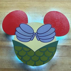 Puede incluir: Una decoración de pared circular de madera con una cara de color amarillo claro, dos orejas rojas grandes, una parte superior en forma de concha morada y un patrón de escamas de pez verde en la mitad inferior. Emite una luz azul suave.