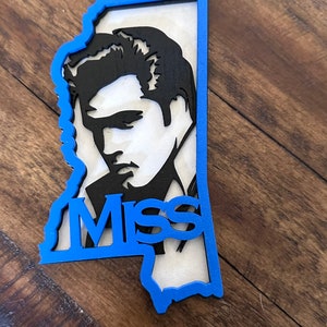 Peut inclure: Une silhouette noire d'Elvis Presley d&eacute;coup&eacute;e dans une forme bleue de l'&eacute;tat du Mississippi. Le mot "Miss" est d&eacute;coup&eacute; dans la forme de l'&eacute;tat.