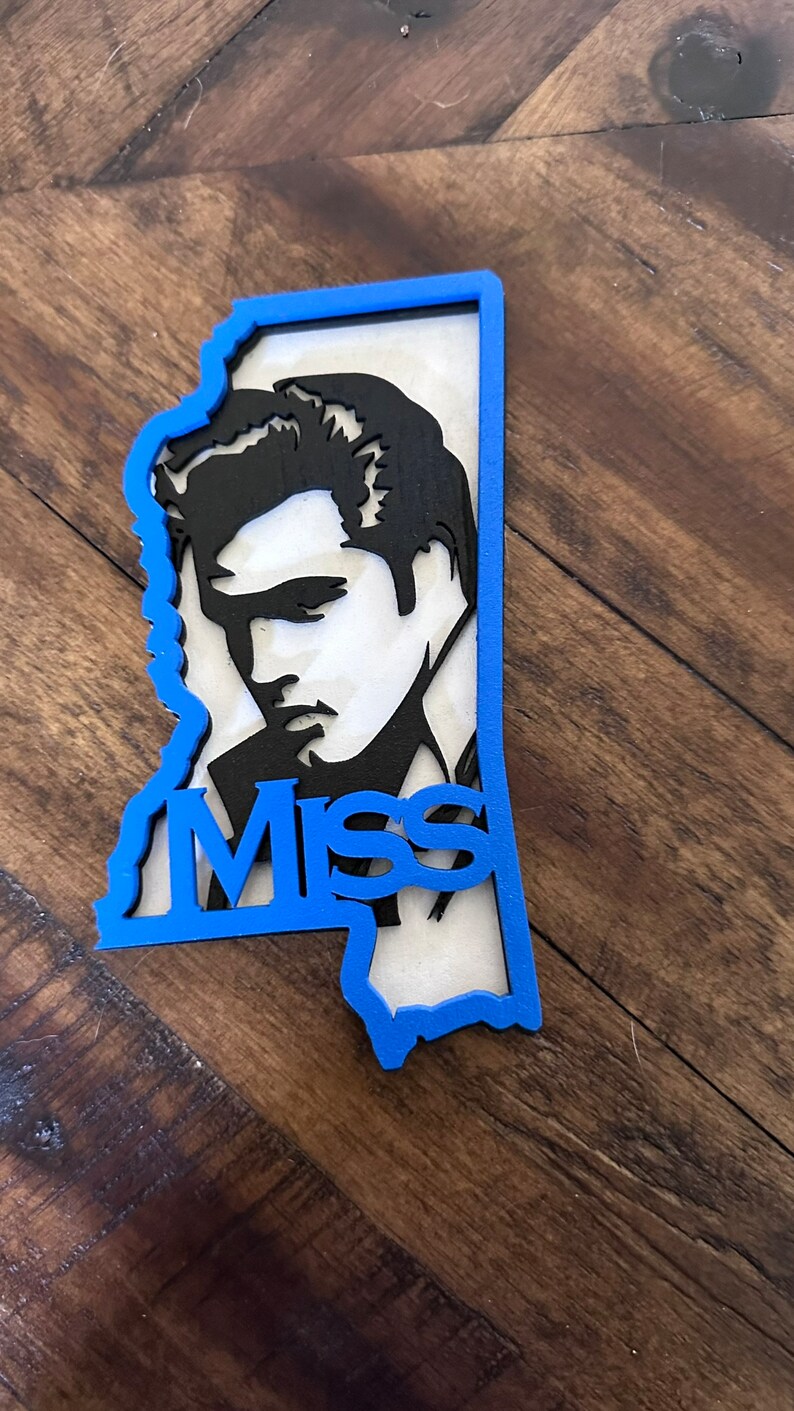 Peut inclure: Une silhouette noire d'Elvis Presley d&eacute;coup&eacute;e dans une forme bleue de l'&eacute;tat du Mississippi. Le mot "Miss" est d&eacute;coup&eacute; dans la forme de l'&eacute;tat.