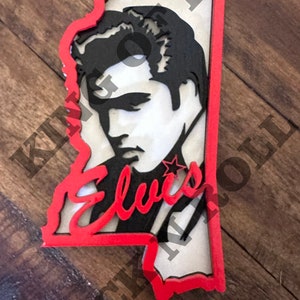 Peut inclure: Une d&eacute;coupe en bois rouge et blanche du visage d'Elvis Presley en forme de l'&eacute;tat du Mississippi. La d&eacute;coupe pr&eacute;sente une image en noir et blanc du visage d'Elvis Presley avec le mot "Elvis" &eacute;crit en rouge sous l'image.