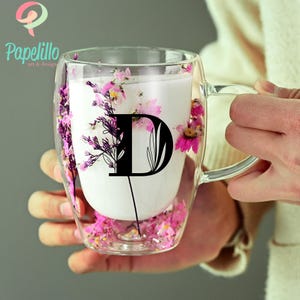 May include: Double-walled glass mug with a handle, filled with a white liquid and decorated with pink flowers. A large black letter "D" is printed on the mug. The mug is held by a person.