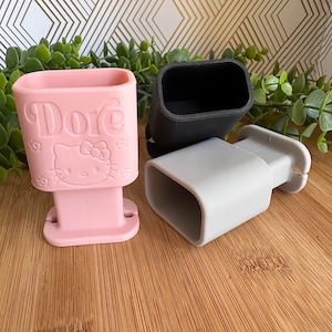 May include: Three silicone phone holders in pink, gray, and black. The pink holder has a Hello Kitty design and the text "Dore" embossed on the front.
