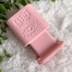 May include: A pink silicone phone stand with a raised eye design and the word "Gigi" embossed on the front.