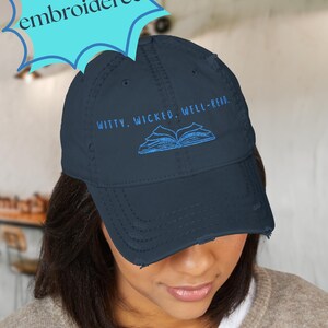 May include: Navy blue distressed baseball cap with the embroidered text "WITTY. WICKED. WELL-READ." and an image of an open book. The word "embroidered!" is in a blue starburst shape above the cap.