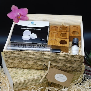Peut inclure: Un coffret en bois contenant des serviettes compressées, de l'huile essentielle et des supports en bois. La boîte est décorée d'un motif floral et d'une étiquette indiquant "Coffret". Une orchidée rose et une petite plante succulente en pot sont également présentes.