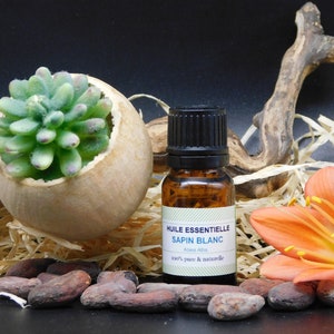 Peut inclure: Une petite bouteille en verre brun avec un bouchon noir contenant de l'huile essentielle. L'étiquette indique "Huile Essentielle Sapin Blanc Abies Alba 100% pure & naturelle". La bouteille est entourée de bois brun, d'herbe sèche et d'une seule fleur orange.