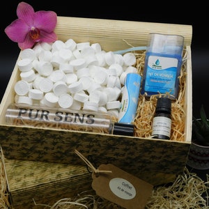 Peut inclure: Un coffret en bois rempli de produits de toilette de voyage, dont une bouteille d'huile essentielle, une petite bouteille de désinfectant pour les mains, un tube de baume à lèvres et un paquet de lingettes nettoyantes pour le visage. Le coffret est étiqueté "Coffret Kit de Voyage".