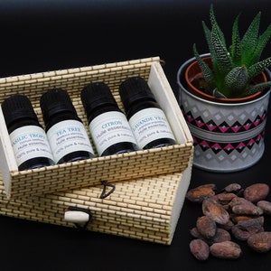 Peut inclure: Un coffret en bois contenant quatre petits flacons en verre d'huiles essentielles. Les flacons sont étiquetés avec les noms des huiles : Basilic Tropical, Tea Tree, Citron et Lavande Vraie. Le coffret est en bois brun clair et présente un motif tissé. Les flacons sont tous en verre transparent et ont des bouchons noirs. Les huiles sont toutes 100% pures et naturelles.