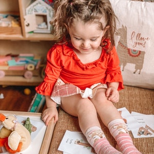 Puede incluir: Una ni&ntilde;a peque&ntilde;a con una camisa roja de manga larga con volantes y una falda estampada. La ni&ntilde;a lleva calcetines a rayas rosas y blancas con caras de ciervos. La ni&ntilde;a est&aacute; sentada en una superficie tejida con tarjetas del alfabeto.
