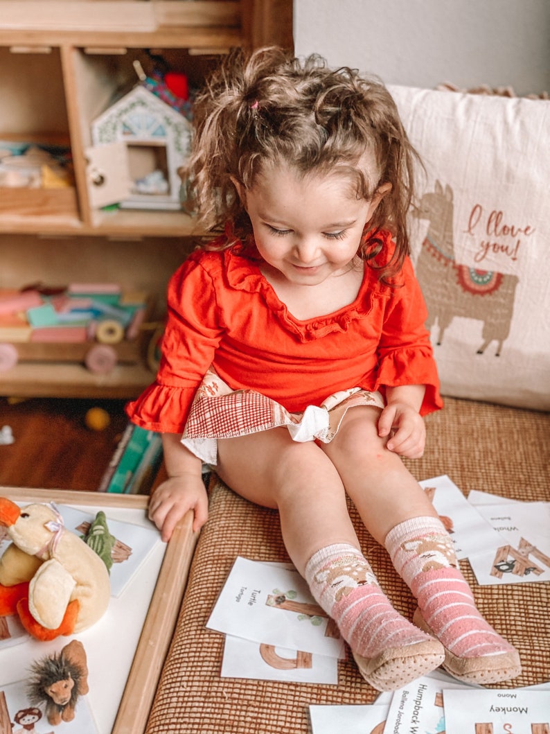 Puede incluir: Una ni&ntilde;a peque&ntilde;a con una camisa roja de manga larga con volantes y una falda estampada. La ni&ntilde;a lleva calcetines a rayas rosas y blancas con caras de ciervos. La ni&ntilde;a est&aacute; sentada en una superficie tejida con tarjetas del alfabeto.