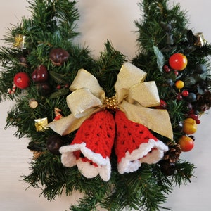 May include: A Christmas wreath decorated with red and green foliage, berries, and a pair of red crocheted bells with white trim. A gold ribbon with a bow is tied around the bells.