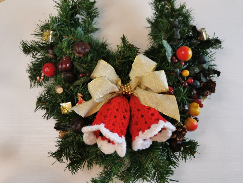 May include: A Christmas wreath decorated with red and green foliage, berries, and a pair of red crocheted bells with white trim. A gold ribbon with a bow is tied around the bells.