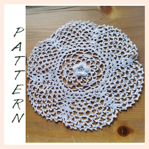 Può includere: Tovaglietta all'uncinetto bianca con un delicato disegno floreale al centro. La tovaglietta è circolare e ha un motivo a pizzo.