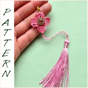May include: A pink crocheted flower charm with a gold clasp and a pink tassel. The charm is attached to a pink cord.