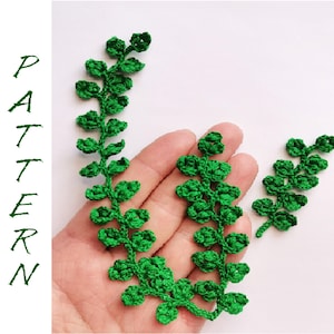 May include: Two crocheted green leafy sprigs, one larger than the other, are displayed on a hand. The sprigs are made of green yarn and have a delicate, intricate design.