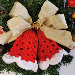 May include: A red and white crocheted Christmas bell ornament with a gold ribbon bow. The ornament is decorated with gold beads and is hanging on a green Christmas wreath.