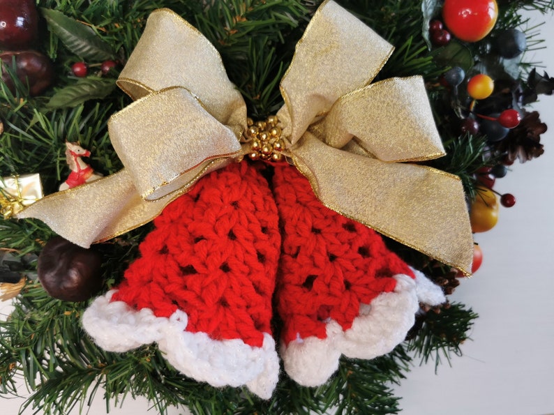 May include: A red and white crocheted Christmas bell ornament with a gold ribbon bow. The ornament is decorated with gold beads and is hanging on a green Christmas wreath.