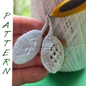 May include: Two white crocheted leaf shapes with a hole in the center of each leaf. The leaves are held in a hand with a white ball of yarn and a yellow crochet hook.