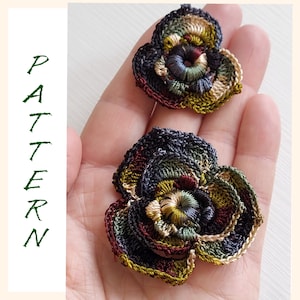 May include: Two crocheted flower embellishments in shades of green, brown, cream, and dark blue. The flowers are layered and have a textured, three-dimensional appearance.