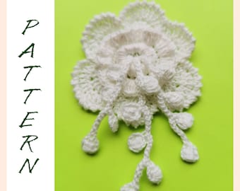 Puede incluir: Flor blanca de ganchillo con un centro fruncido y bucles colgantes. La flor está sobre un fondo verde.
