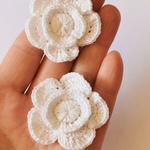 May include: Two white crocheted flowers with a layered design. The flowers are made with a delicate white yarn and have a simple, elegant look.