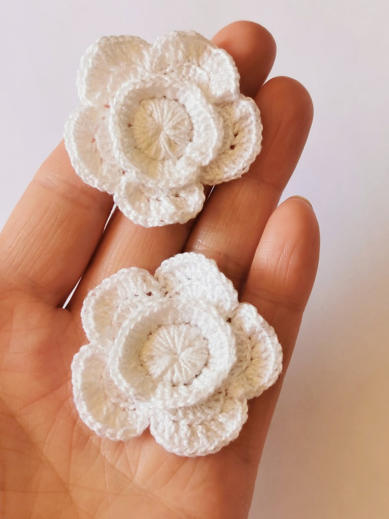 May include: Two white crocheted flowers with a layered design. The flowers are made with a delicate white yarn and have a simple, elegant look.