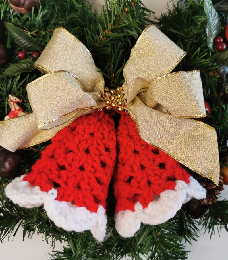 May include: A red crocheted bell ornament with white trim, tied with a gold ribbon bow, on a green Christmas wreath.