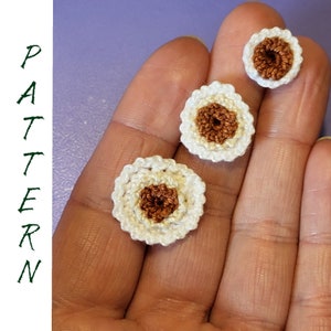 May include: Three crocheted white flowers with brown centers. The flowers are different sizes and are arranged in a row on a hand.