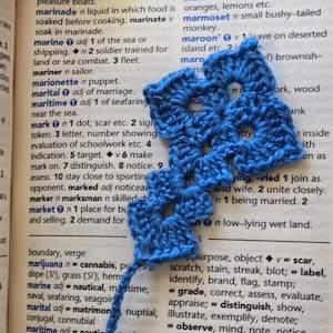 Pode incluir: Um marcador de livro de crochê azul feito à mão com um design floral. O marcador de livro é feito com um ponto texturizado, e o padrão vazado é visível. O marcador de livro está colocado em uma página de livro aberta com entradas de dicionário.
