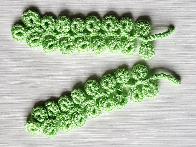 May include: Two crocheted green leafy sprigs with a delicate, intricate design. The sprigs are made of yarn and have a natural, organic look.