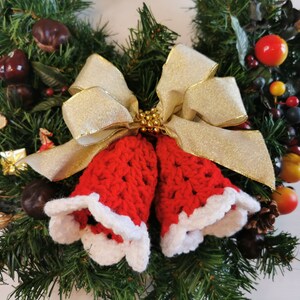May include: A Christmas wreath decorated with red and white crocheted bells, a gold bow, and various greenery and berries.