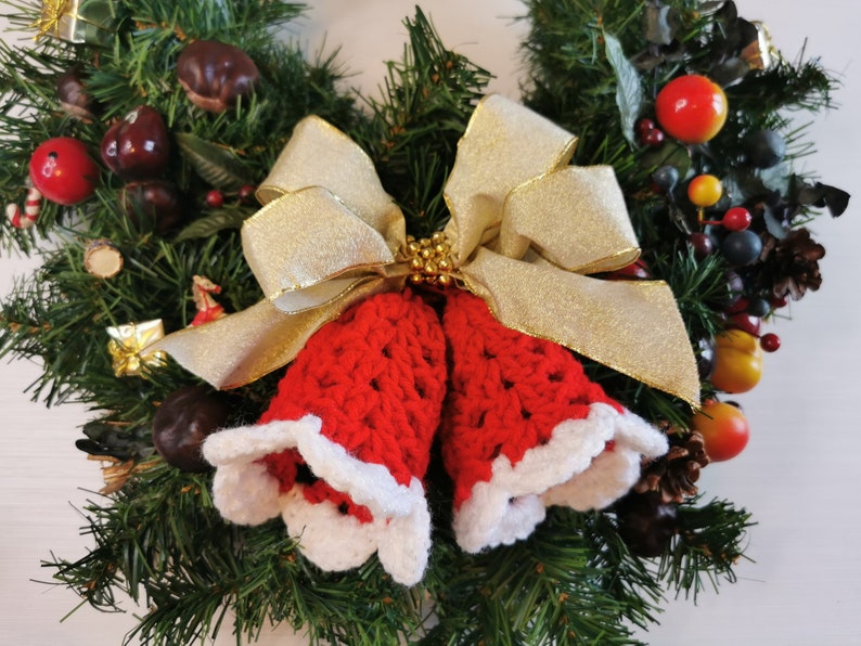 May include: A Christmas wreath decorated with red and white crocheted bells, a gold bow, and various greenery and berries.
