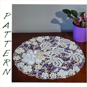 May include: White crochet doily with a large white flower in the centre and purple crocheted accents. The doily is on a wooden table.