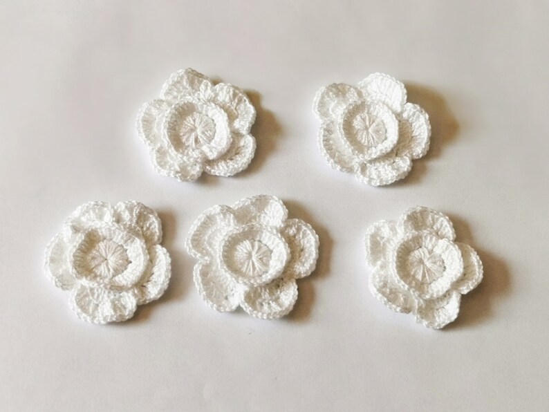 May include: Five white crocheted flowers with a layered design. The flowers are arranged in a semi-circle on a white background.