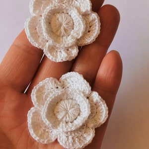 May include: Two white crocheted flowers with a layered design. The flowers are made with a delicate white yarn and have a simple, elegant look.