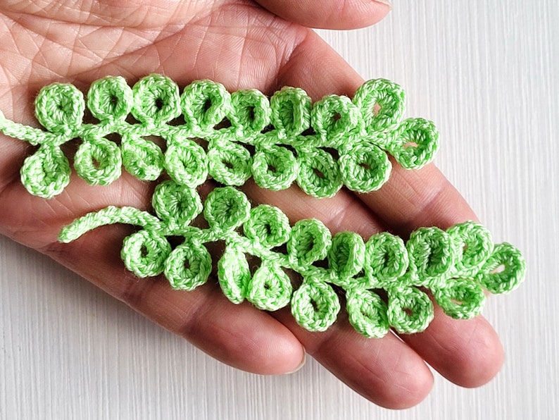 May include: Two green crocheted leaf sprigs with a delicate, lacy design. Each sprig has multiple small, rounded leaves.